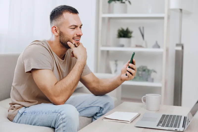 Smiling man using smartphone while sitting on a couch, wearing casual clothes, relaxed atmosphere, modern interior design, engaging with a digital device.