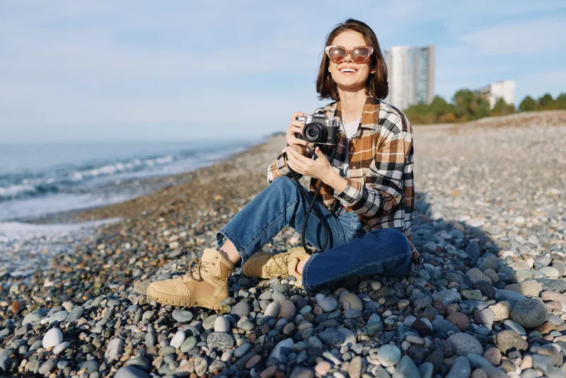 Woman photographer sits on a pebble beach, smiling behind a vintage camera, wearing a plaid shirt and boots, capturing relaxed outdoor seaside moments with focused creativity, with bright energy.