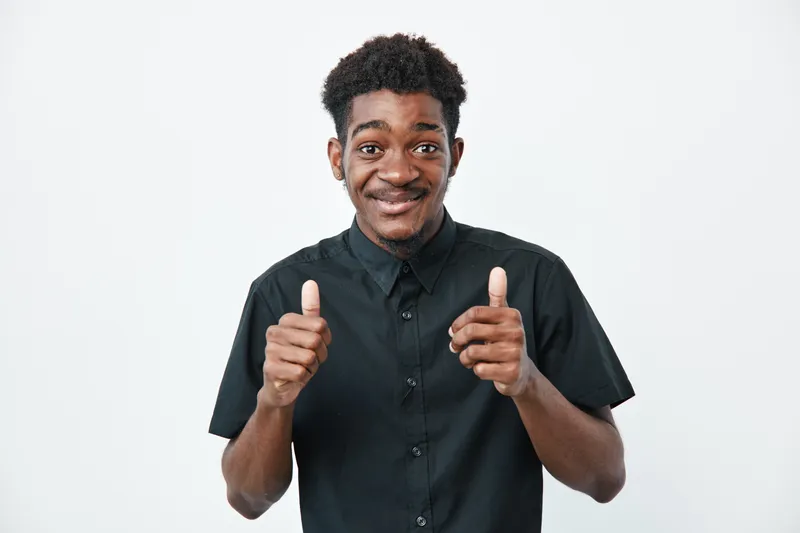 Young black man with dark skin wearing black shirt showing thumbs up gesture smiling confidently on white background. Positive expression and casual style for success or approval.