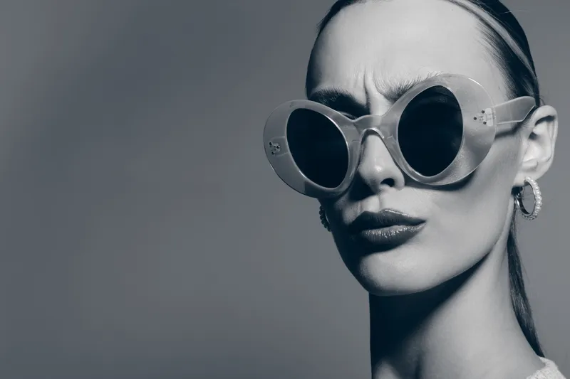 Close-up portrait of a young white woman with pale skin wearing large round sunglasses and silver hoop earrings. Studio shot with dramatic lighting and serious facial expression.