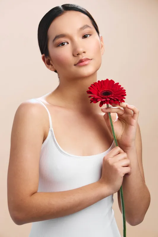 asian woman holds a bright red flower near her face, wearing a light tank top, soft lighting, serene expression, and graceful posture conveying calm beauty and elegance.