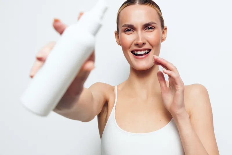 Smiling woman with clear skin holding white bottle close to camera on white background, skincare concept, beauty routine, natural makeup, wellness and health care.