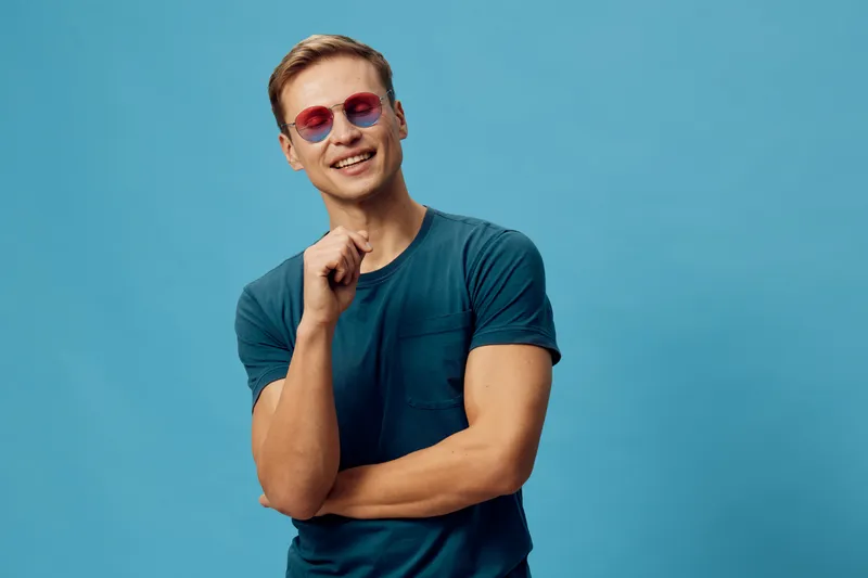 Smiling young caucasian man with short brown hair wearing pink sunglasses and blue t-shirt, posing confidently with hand near face on plain blue background, studio portrait. People lifestyle concept