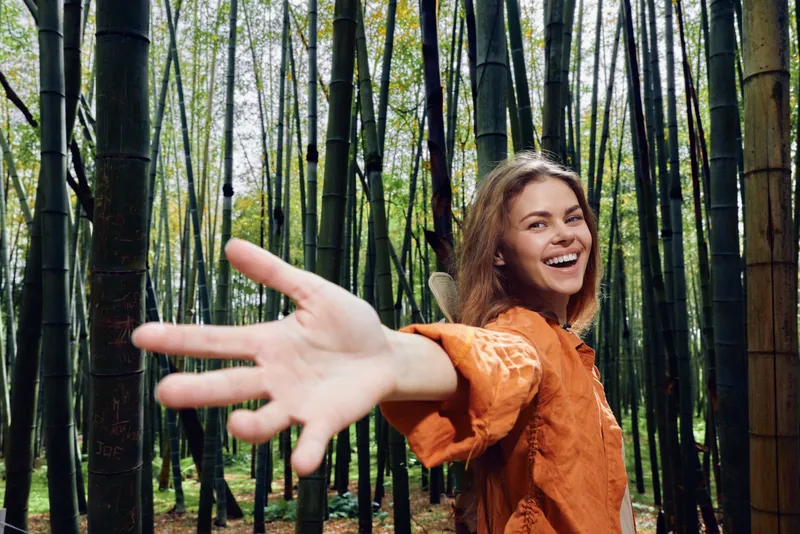 Woman smiling in a bamboo forest reaching hand toward the camera, portrait of a joyful young person in nature wearing an orange top, playful outdoor mood and travel lifestyle.