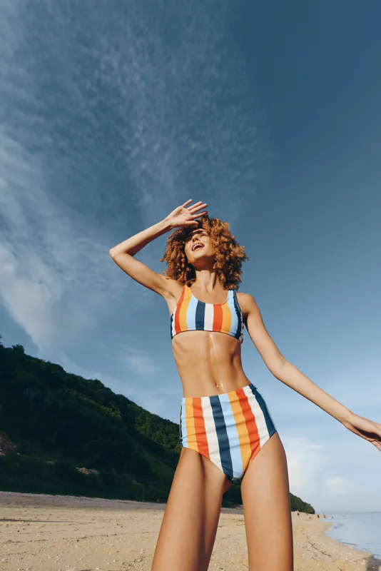 Young woman in a striped bikini stands on a sunlit beach, shielding her eyes with one hand as the ocean breeze lifts her hair, capturing a vibrant, carefree summer moment.