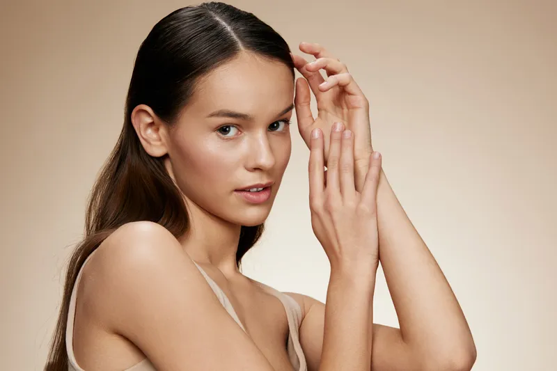 Young woman with long hair holding her hands near her face, showcasing natural beauty, soft lighting, neutral background, ideal for wellness and skincare themes
