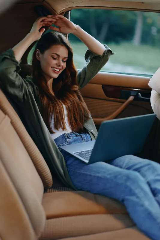 Relaxed young woman enjoying her time while working on a laptop in a car, dressed casually in a white top and jeans, with a cheerful and content expression in natural surroundings.