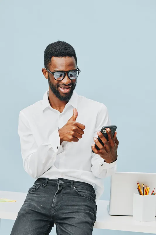 Confident young African American man with glasses wearing white shirt holding smartphone and smiling in modern office setting. People technology concept, casual style, positive mood, workspace environ