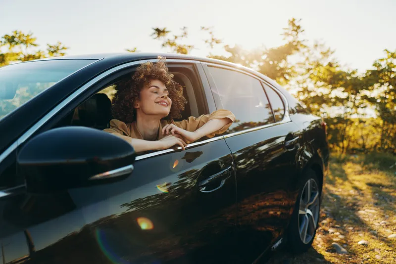 A joyful woman leans out of a car window, hair in the breeze, basking in warm sunlight. A candid lifestyle moment with retro film color and carefree vibe.