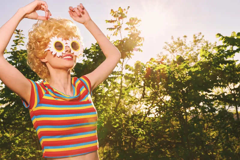 A woman enjoys outdoor sunny moments in a colorful striped top, wearing playful flower sunglasses. A retro lifestyle moment captured with bright film tones and a carefree mood.