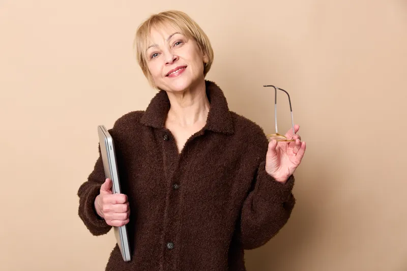 Elderly woman portrait, smiling indoors, holding glasses and smartphone, brown fleece jacket, beige background, calm positive mood, senior, lifestyle, technology