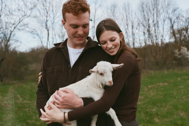 Young couple embraces outdoors as the woman holds a white baby goat in her arms, sharing a tender moment in a natural spring setting with bare trees in the background.