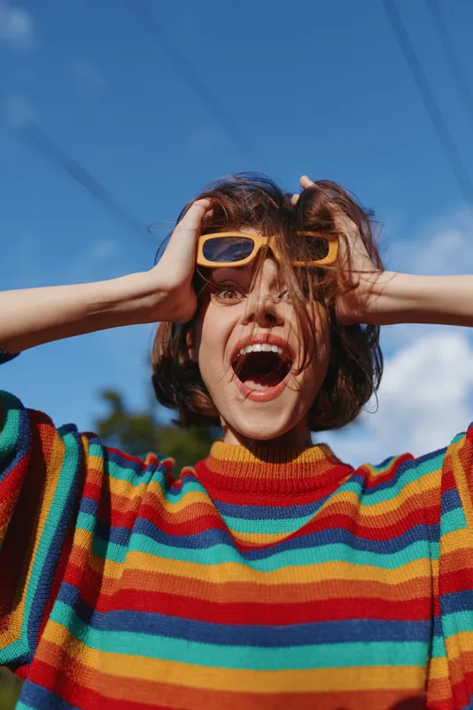 Woman portrait in a colorful sweater with sunglasses shouting joyfully outdoors, fashion look and energetic expression, rainbow stripes, short hair and sunny blue sky backdrop.