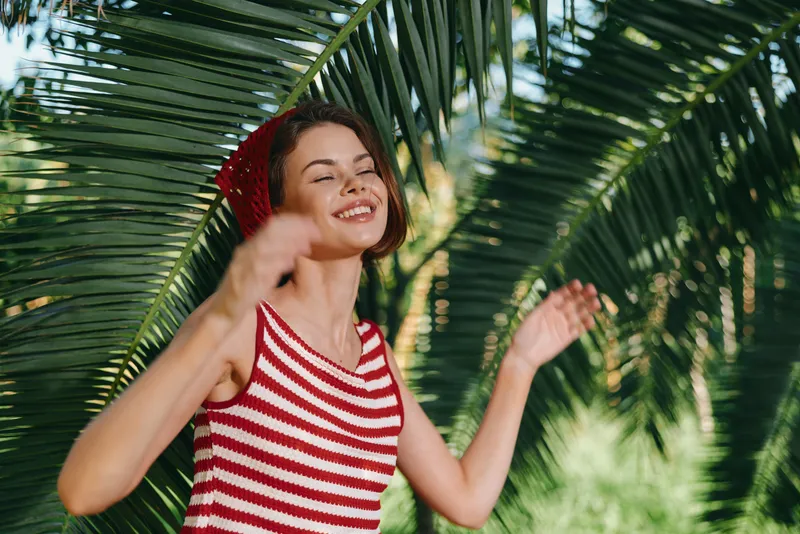 A cheerful woman in a striped red and white top stands under palm leaves, smiling and enjoying a sunny moment outdoors. Bright natural light adds a lively, carefree mood.