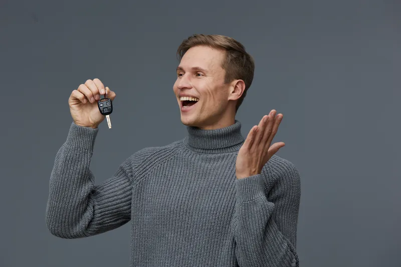 Smiling man holding car key with happy expression, wearing casual gray sweater, against plain gray background. Portrait of a joyful person celebrating new vehicle purchase or achievement, lifestyle co
