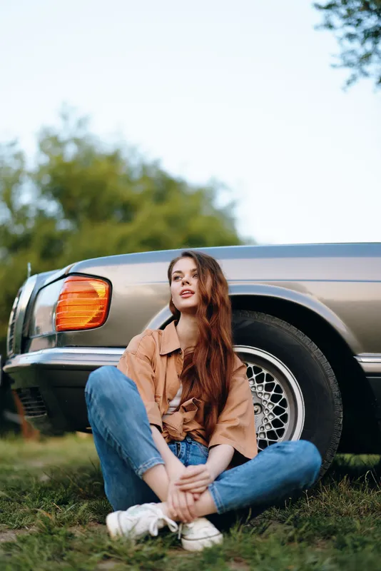 Young woman with long hair wearing a stylish outfit sitting by a vintage car on a sunny day, embodying a relaxed lifestyle and nostalgia.
