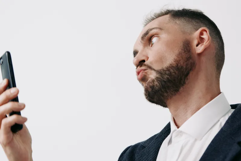 A professional man in business attire stands against an isolated colored background, showing a thoughtful, contemplative emotion while confidently checking a smartphone during a work moment.