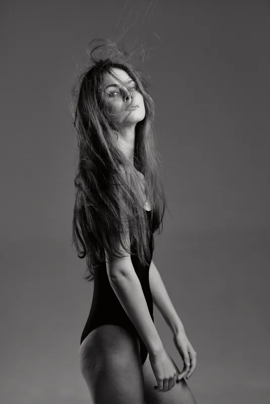 Black and white portrait of a young woman with long hair wearing a black bodysuit posing with an elegant serious expression