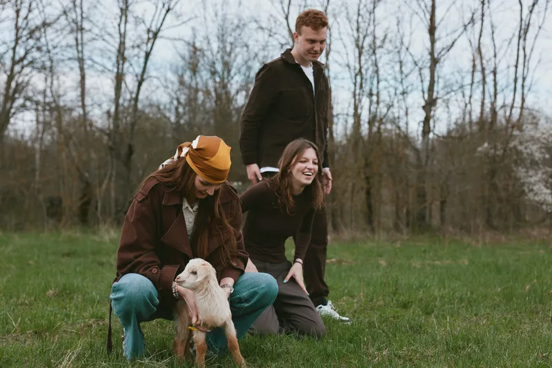 A cheerful family outdoors with a gentle goat kid in a grassy field, capturing a candid moment of bonding and play. A warm lifestyle scene in a rural setting.