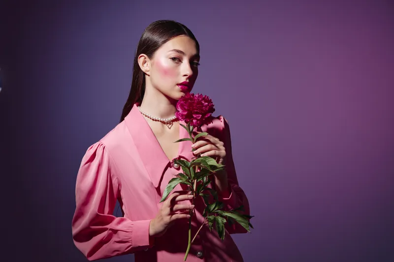Young woman in pink suit holding peony flower with soft makeup. Creative beauty fashion portrait on purple background. Concept for elegant style and floral inspiration.