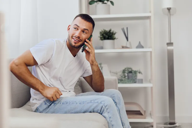 Smiling man on a sofa, engaged in a phone conversation, exuding warmth and comfort, surrounded by modern decor in a cozy living room setting.