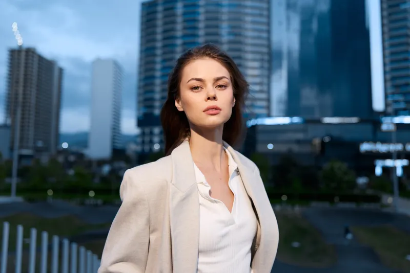 Confident woman with serious expression in business attire outdoors at twilight in urban financial district with modern skyscrapers