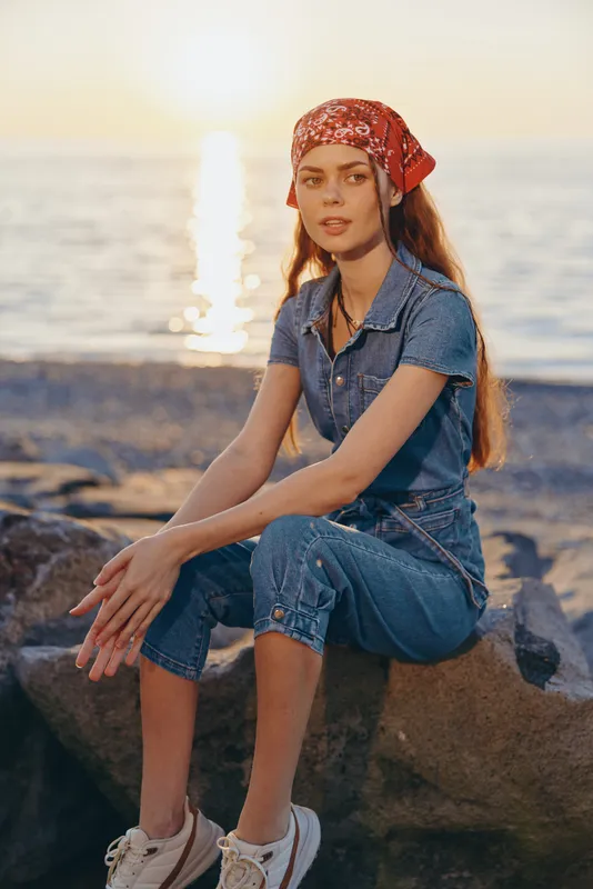 Lifestyle woman in modern boho-western denim street style wearing a red bandana, sitting by the beach at sunset with warm film color tones and relaxed mood.