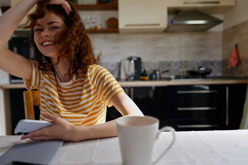 Young woman with striped shirt sits at a bright kitchen table, checking a smartphone and smiling warmly, while a white mug sits in the foreground and soft light fills the room.