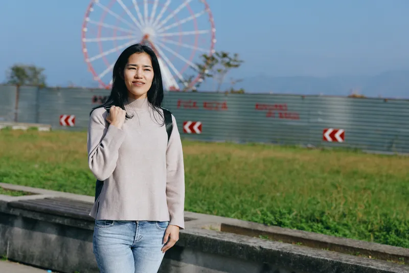 Woman posing outdoors near a ferris wheel, casual sweater and jeans, bright day, open space, amusement backdrop, relaxed style, candid moment, travel vibe, scenic park setting