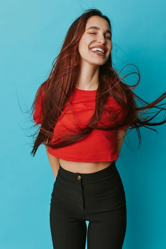 Young woman with long hair smiling with closed eyes wearing red crop top and black pants standing against blue background. Happy female model with casual style and natural expression.