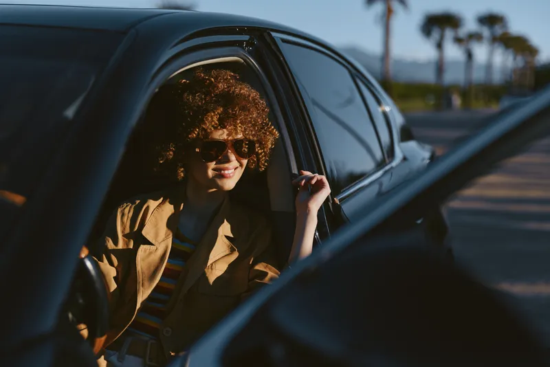 Casual woman with curly hair sits in a car, wearing a jacket, sunlight highlights retro tones, a lifestyle portrait with a calm mood and warm outdoor glow captured through window glare