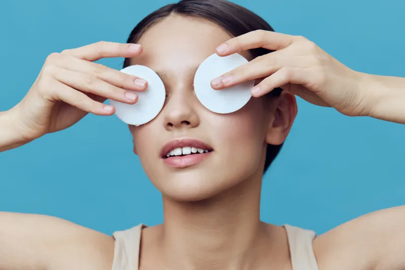 Young woman relaxing with cotton pads over her eyes, showcasing skincare routine with a serene expression on a vibrant blue background
