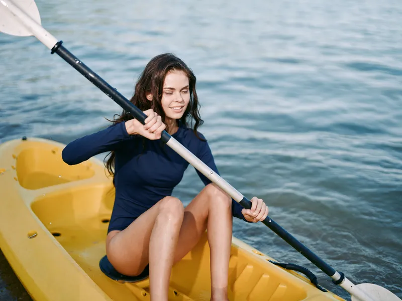 A young woman paddles a yellow kayak on calm water, gripping a double-blade paddle, smiling as she rows, capturing outdoor summer adventure and bright sunlight.