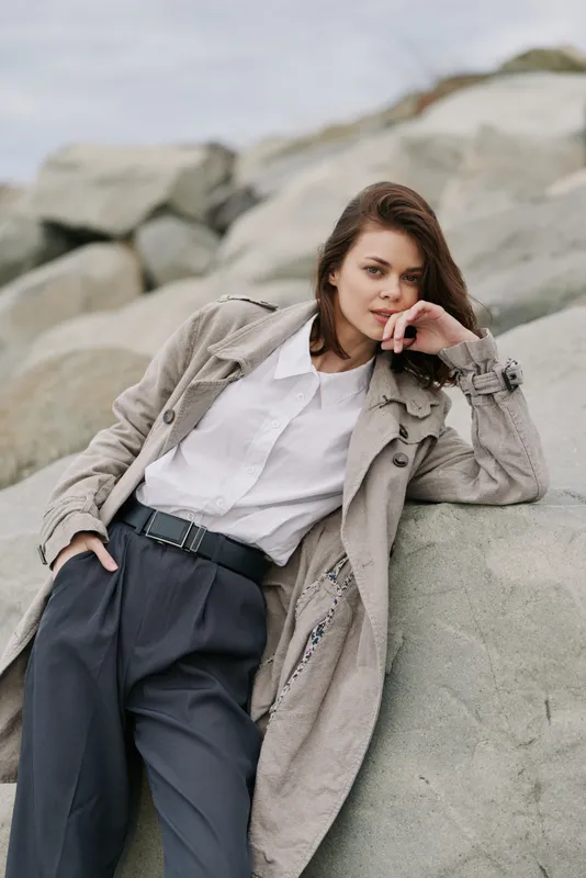 Young woman in stylish coat and white shirt leans on rocks outdoors. Casual fashion, natural makeup, and relaxed mood in a serene outdoor setting with neutral tones.