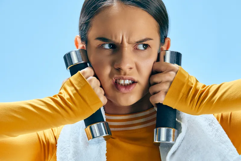 Fitness woman in bright sportswear grips dumbbells, intense expression, mid workout, gym vibe with energetic focus and determination during training.