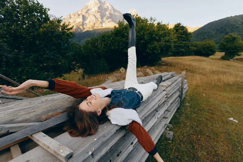 Woman lies relaxed on a wooden fence in a meadow beneath a mountain, outdoors enjoying nature and relaxation; casual clothing and boots, smiling and resting in scenic countryside.
