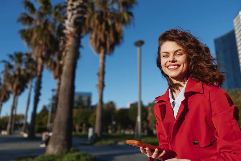 Cheerful woman outdoors in a red coat, smiling under bright sun, holding a phone, palm trees and blue sky behind, casual urban park moment in warm light
