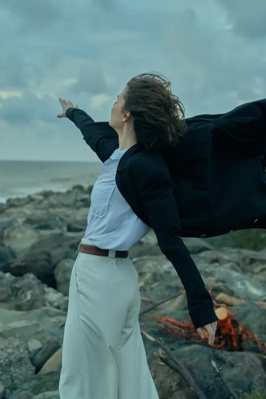 Windblown cape and dress flow as a fashion forward figure stands on rocky coastline, outdoors by the ocean, conveying freedom and dramatic motion