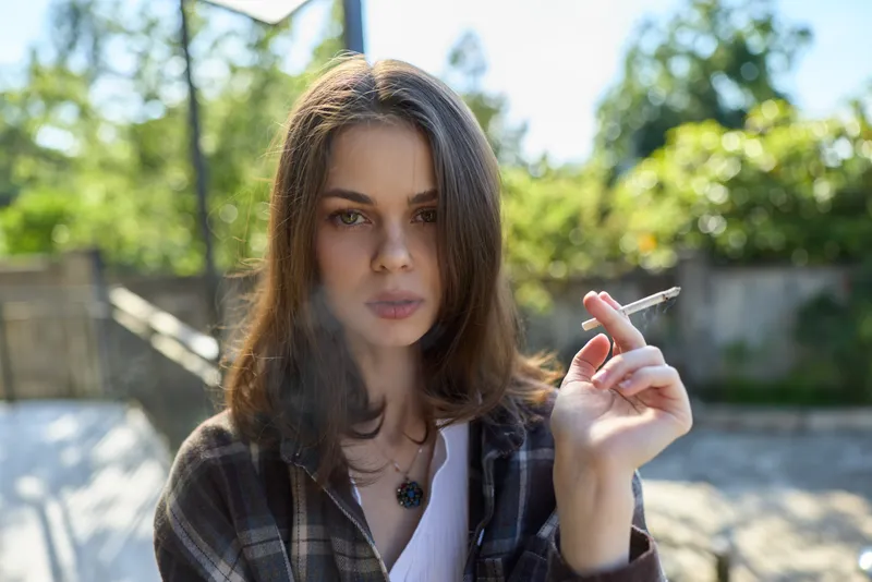 Young woman with shoulder length brunette hair relaxes outdoors, cigarette in hand, sunlit moment with soft breeze and lush background