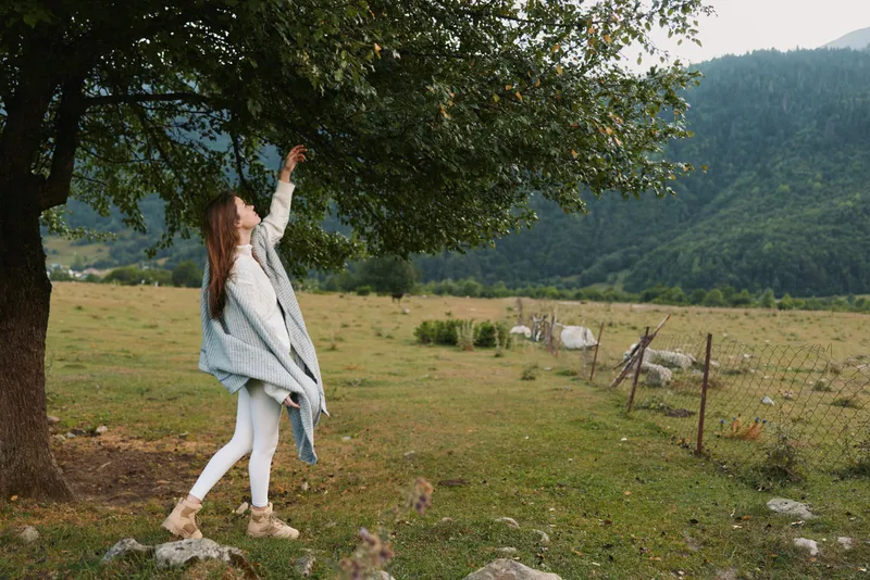 Young woman in white leggings and beige boots reaches up to touch leaves on a large tree in a rural field with mountains in the background.