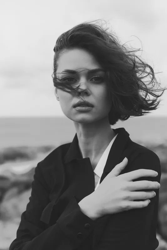 Moody black and white image of a woman in a blazer with windblown hair and sunglasses outdoors by the sea