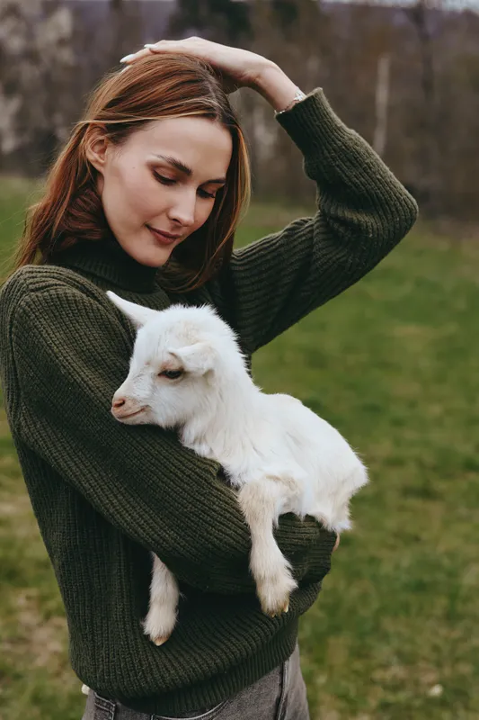 Cheerful woman outdoors, gently holding a white goat kid, sunny day, cozy forest backdrop, casual sweater, tender moment, natural colors, calm and warm mood