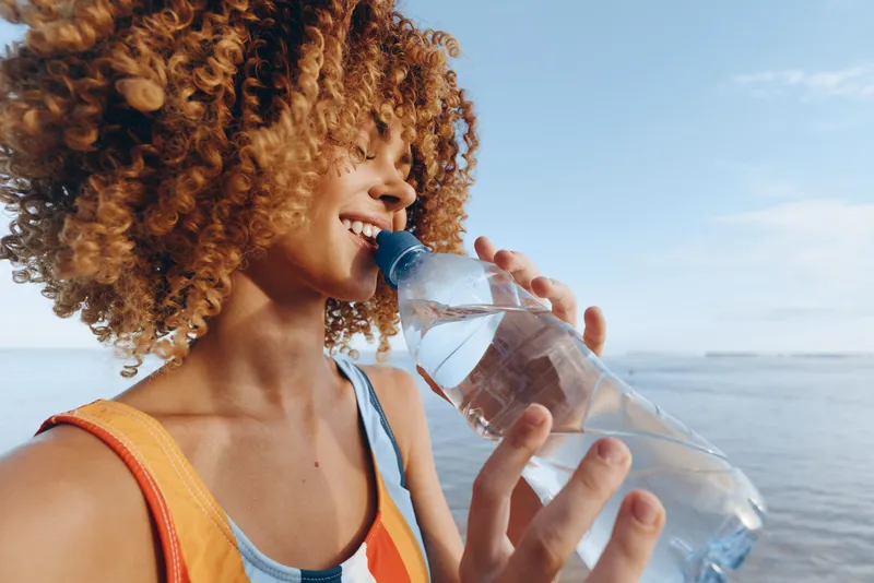 Woman drinking water, curly hair, smiling outdoors by sea in summer, orange backpack straps visible, closeup on hydration, refreshing healthy lifestyle, natural light, calm horizon.