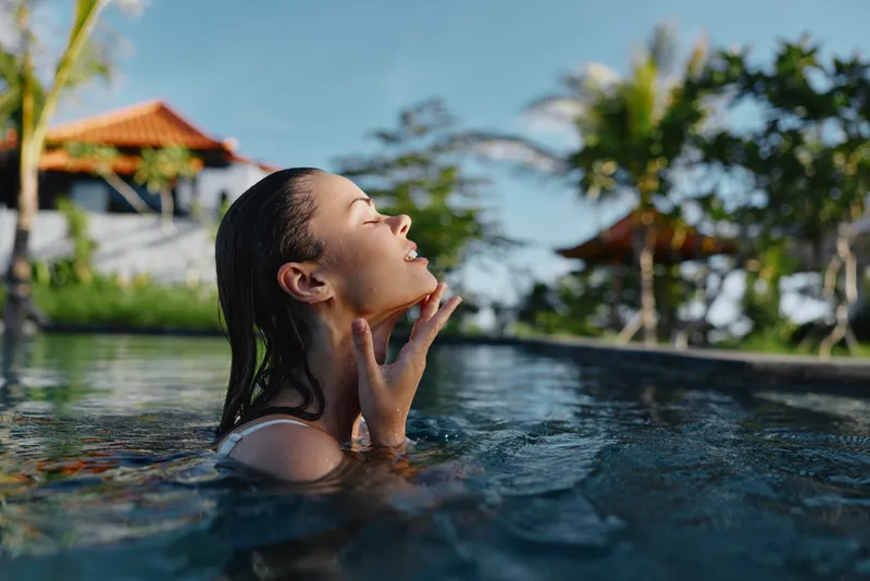 Relaxed pool scene with a woman emerging from clear water, eyes closed, hands near her face, sunny tropical setting, outdoor resort mood, serene moment of leisure