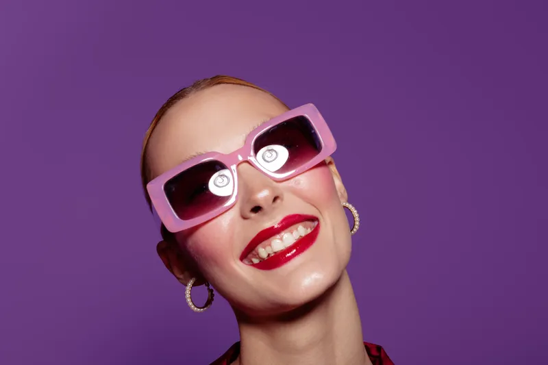 Smiling woman with bright red lipstick wears large pink sunglasses and hoop earrings against purple background. Concept of fashion, beauty, and confident style portrait.
