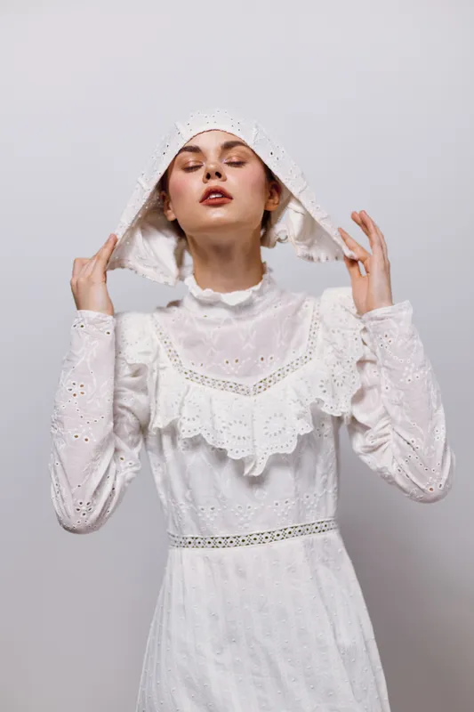 Elegant young woman in a white vintage dress and head covering, expressing serenity and confidence against a neutral background
