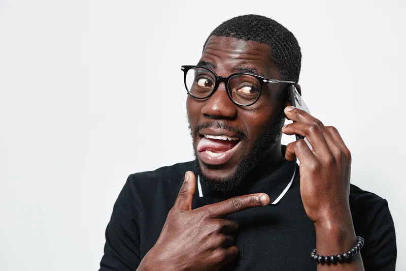 African American man with dark skin wearing glasses talking on smartphone and pointing finger at phone with playful expression on white background in studio.