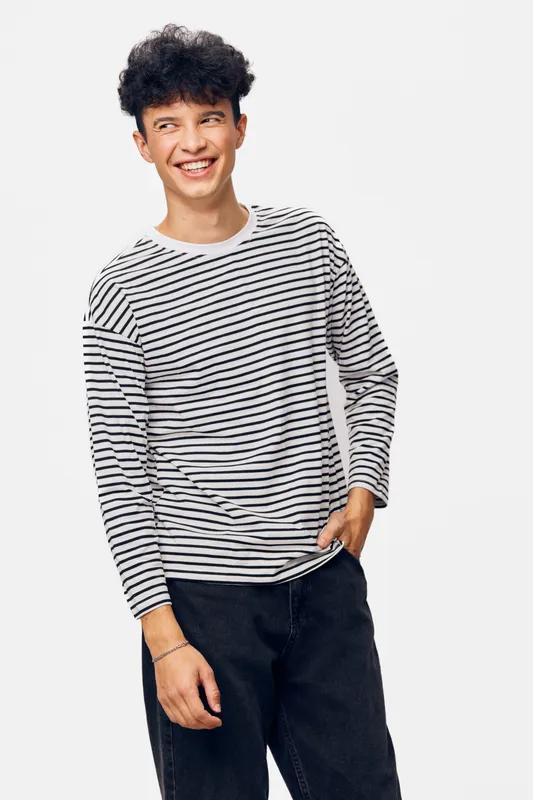 Young man smiling wearing a striped long-sleeve shirt and black pants against a plain white background, expressing casual and relaxed mood.