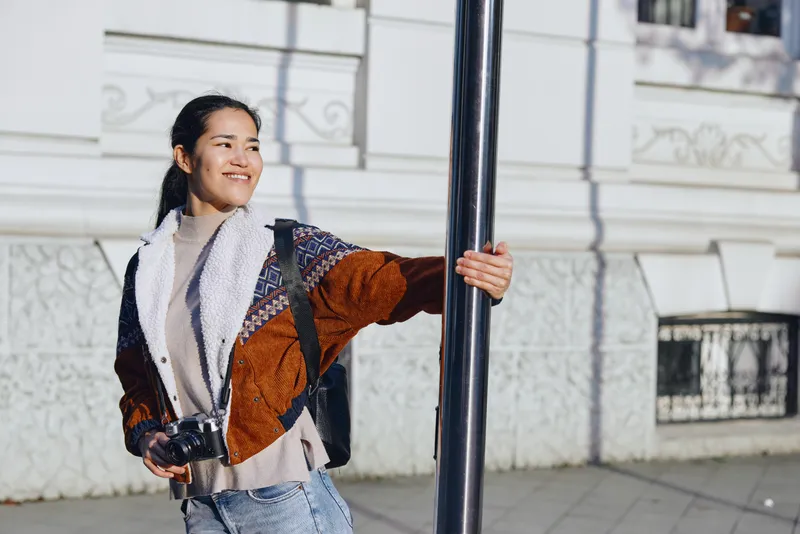 Woman with camera smiling beside a street pole in an outdoor urban setting, casual fashion coat and scarf, candid street style mood, daylight photography vibe, cityscape backdrop, ready to capture