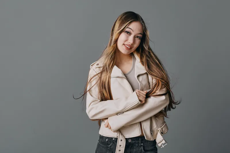 Young woman with long hair wearing light jacket and black jeans posing with crossed arms and smiling on gray background in studio portrait.